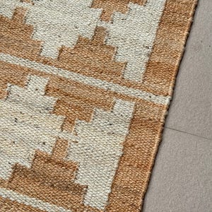 May include: A woven rug featuring a repeating geometric pattern in tan and off-white. The texture is coarse and fibrous, with a distinct grid-like weave. The design consists of stepped diamond shapes arranged in horizontal rows across the surface.