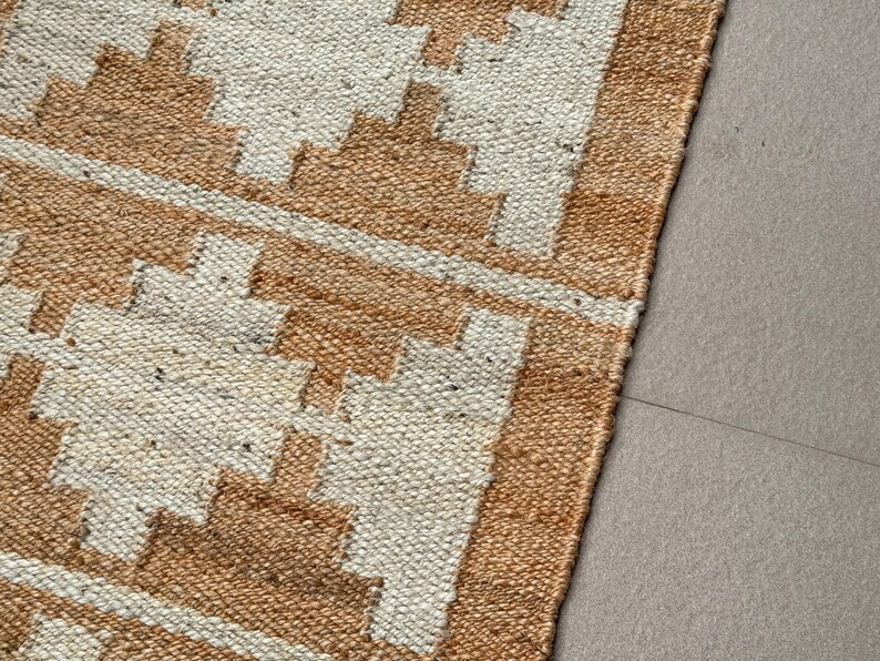 May include: A woven rug featuring a repeating geometric pattern in tan and off-white. The texture is coarse and fibrous, with a distinct grid-like weave. The design consists of stepped diamond shapes arranged in horizontal rows across the surface.
