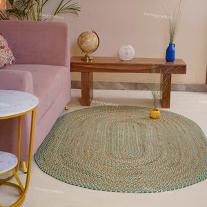 Puede incluir: Alfombra ovalada trenzada en tonos verdes, beige y naranja. La alfombra est&aacute; sobre un suelo de color claro, con un sof&aacute; rosa y una mesa de madera al fondo. Una peque&ntilde;a mesa auxiliar con tapa de m&aacute;rmol est&aacute; en primer plano.