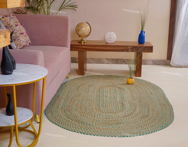 Puede incluir: Alfombra ovalada trenzada en tonos verdes, beige y naranja. La alfombra est&aacute; sobre un suelo de color claro, con un sof&aacute; rosa y una mesa de madera al fondo. Una peque&ntilde;a mesa auxiliar con tapa de m&aacute;rmol est&aacute; en primer plano.