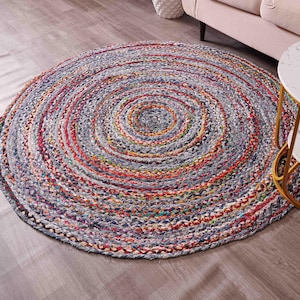 K&ouml;nnte beinhalten: Ein runder geflochtener Teppich mit Spiralmuster. Der Teppich weist eine Mischung aus Farben auf, darunter Grau, Rot, Orange und Blau. Er liegt auf einem Holzboden, im Hintergrund sind ein Sofa und ein Beistelltisch zu sehen.