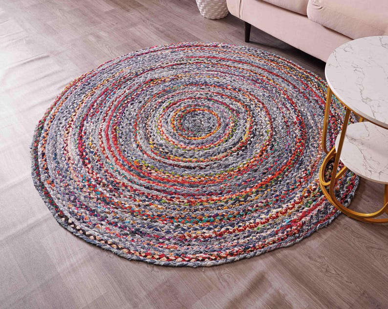 K&ouml;nnte beinhalten: Ein runder geflochtener Teppich mit Spiralmuster. Der Teppich weist eine Mischung aus Farben auf, darunter Grau, Rot, Orange und Blau. Er liegt auf einem Holzboden, im Hintergrund sind ein Sofa und ein Beistelltisch zu sehen.