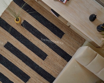 May include: A rectangular jute rug with a natural tan background and bold black diagonal stripes. The rug is placed on a light-colored wooden surface, with a small yellow vase and decorative objects nearby. The text "THEINDOORRUGS" is visible.
