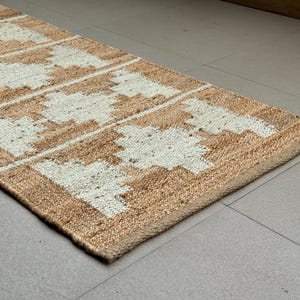May include: A rectangular runner rug featuring a natural tan jute base with a repeating white geometric pattern. The woven texture is visible throughout the surface, showcasing a series of symmetrical, stepped diamond shapes arranged in a linear row.