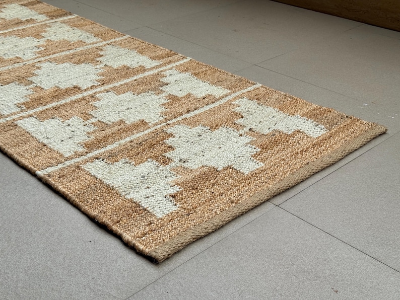 May include: A rectangular runner rug featuring a natural tan jute base with a repeating white geometric pattern. The woven texture is visible throughout the surface, showcasing a series of symmetrical, stepped diamond shapes arranged in a linear row.