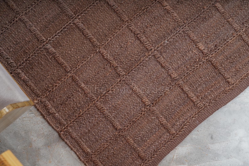 May include: A dark brown woven rug featuring a raised grid pattern of squares. The texture is dense and uniform, created by thick, interlaced fibers that form a consistent geometric design across the entire surface of the rectangular floor covering.