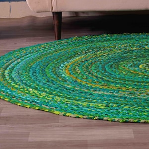 K&ouml;nnte beinhalten: Ein runder geflochtener Teppich in Gr&uuml;n-, T&uuml;rkis- und Gelbt&ouml;nen. Der Teppich besteht aus einem dicken, strukturierten Material und hat ein Spiraldesign. Er liegt auf einem Holzboden, mit einem wei&szlig;en Korb und dem Bein eines beigen Sofas im Hintergrund.