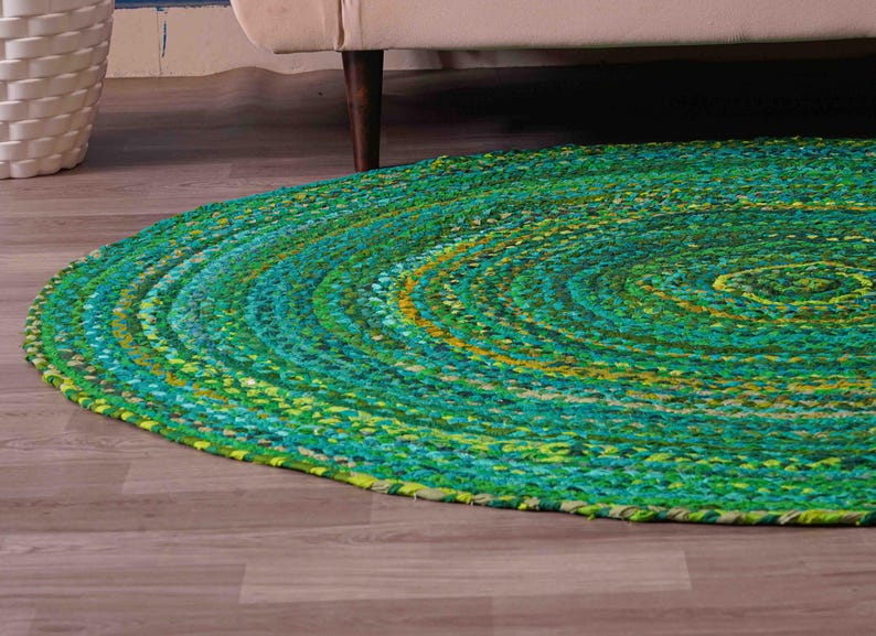 K&ouml;nnte beinhalten: Ein runder geflochtener Teppich in Gr&uuml;n-, T&uuml;rkis- und Gelbt&ouml;nen. Der Teppich besteht aus einem dicken, strukturierten Material und hat ein Spiraldesign. Er liegt auf einem Holzboden, mit einem wei&szlig;en Korb und dem Bein eines beigen Sofas im Hintergrund.