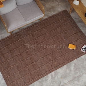 May include: A rectangular brown area rug featuring a raised grid pattern across its surface. The rug displays the text Theindoorrugs printed in the center. The design consists of a repeating square texture throughout the entire piece of floor covering.