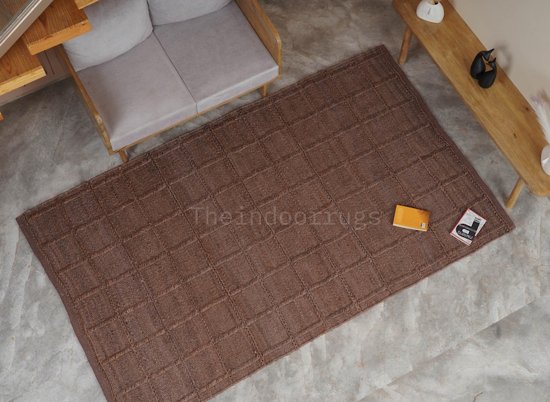 May include: A rectangular brown area rug featuring a raised grid pattern across its surface. The rug displays the text Theindoorrugs printed in the center. The design consists of a repeating square texture throughout the entire piece of floor covering.