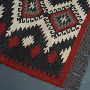 May include: A geometric patterned rug with a red border and fringe. The rug features a repeating pattern in black, white, and red. The rug is made of woven material and has a textured appearance.