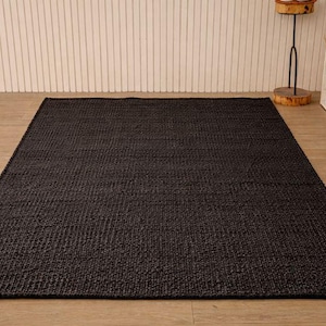 May include: A dark charcoal-coloured rectangular rug with a woven texture. The rug is placed on a light-coloured wooden floor. The background features a white wall with vertical lines.