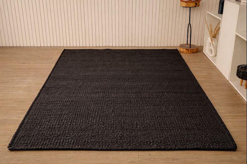 May include: A dark charcoal-coloured rectangular rug with a woven texture. The rug is placed on a light-coloured wooden floor. The background features a white wall with vertical lines.