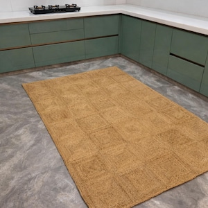 May include: A rectangular, woven jute rug with a geometric pattern. The rug is a warm tan color and is placed on a gray concrete floor. The rug is in a kitchen setting with green cabinets and a white countertop.