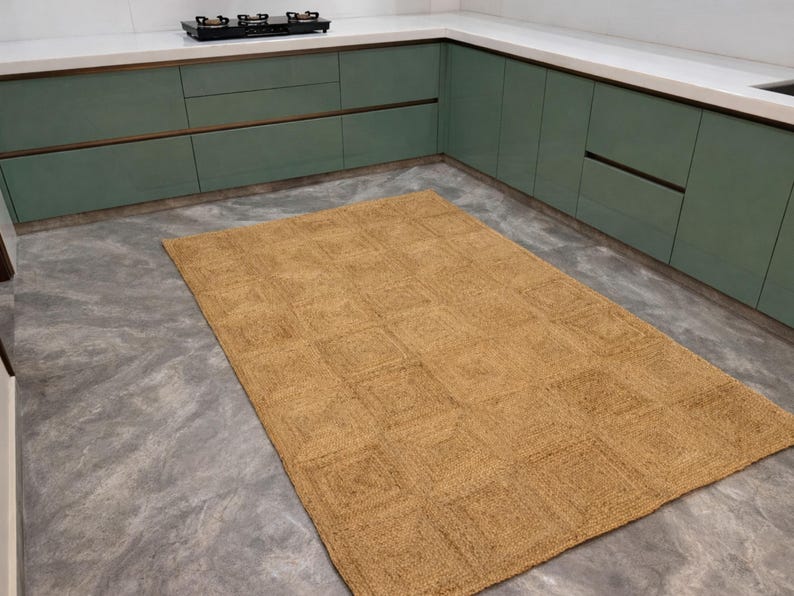 May include: A rectangular, woven jute rug with a geometric pattern. The rug is a warm tan color and is placed on a gray concrete floor. The rug is in a kitchen setting with green cabinets and a white countertop.
