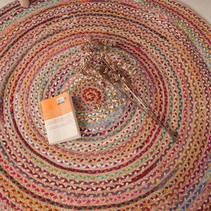 Puede incluir: Una alfombra redonda de yute trenzado con un diseño en espiral en tonos beige, naranja, rosa y verde. Un pequeño libro y flores secas descansan sobre la alfombra. El diámetro de la alfombra es de aproximadamente 122 cm.