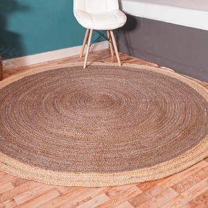 Könnte beinhalten: Ein runder Jute-Teppich mit natürlicher Faseroptik. Der Teppich hat ein konzentrisches Design mit einem helleren Rand und einer dunkleren Mitte. Der Teppich liegt auf einem Holzboden, mit einem weißen Stuhl und dekorativen Gegenständen im Hintergrund.