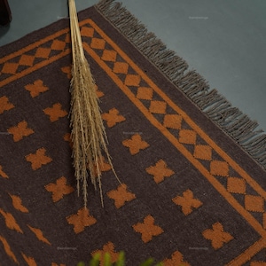 May include: A rectangular rug featuring a dark brown base with a repeating orange cross pattern and a diamond-shaped border. The rug is finished with a fringe along the edge and is decorated with a bundle of dried wheat stalks placed on top.