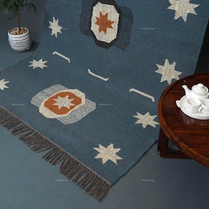 May include: A blue rug with a star pattern and fringe. The rug features star and geometric designs in white, orange, and gray. The rug is displayed on a gray surface, with a small plant and a wooden table with a tea set in the background.