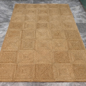 May include: A rectangular jute rug with a geometric square pattern. The rug is a natural tan color and appears to be made of woven fibers. The rug is placed on a gray surface.