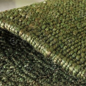Può includere: Primo piano di un tappeto verde tessuto con una superficie strutturata. Il tappeto è realizzato con fibre spesse e intrecciate, creando un aspetto denso e resistente. Il colore è un verde intenso e terroso, con variazioni di tonalità che aggiungono profondità. La costruzione del tappeto suggerisce un materiale in fibra naturale.