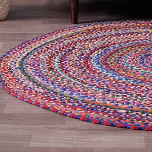 K&ouml;nnte beinhalten: Ein runder, geflochtener Teppich mit einer lebendigen Mischung aus Farben wie Rot, Blau, Rosa und Gelb. Das Design des Teppichs zeigt konzentrische Kreise und erzeugt eine strukturierte Oberfl&auml;che. Der Teppich liegt auf einem Holzboden.