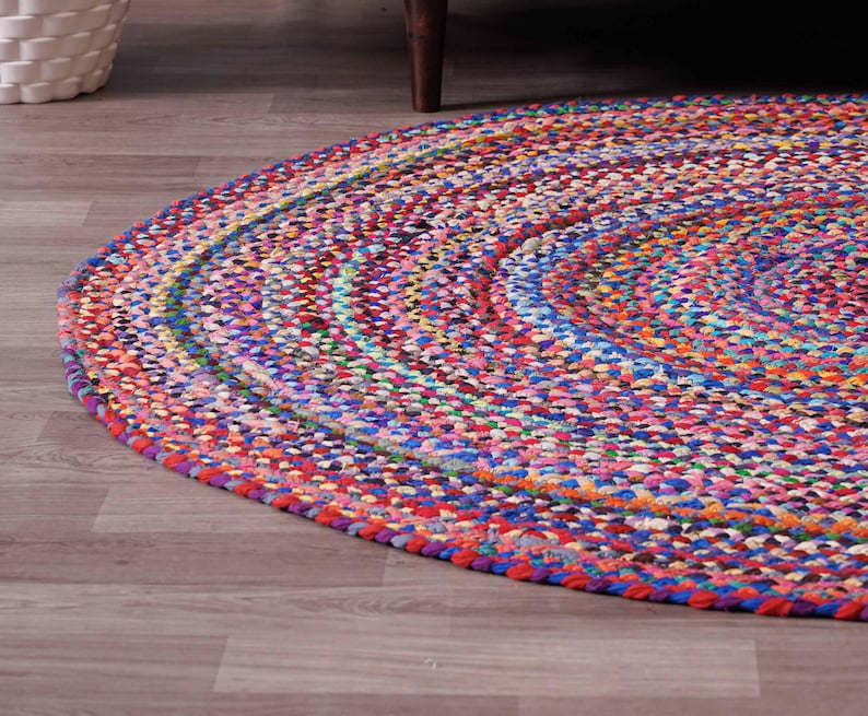 K&ouml;nnte beinhalten: Ein runder, geflochtener Teppich mit einer lebendigen Mischung aus Farben wie Rot, Blau, Rosa und Gelb. Das Design des Teppichs zeigt konzentrische Kreise und erzeugt eine strukturierte Oberfl&auml;che. Der Teppich liegt auf einem Holzboden.