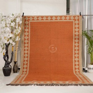 May include: An orange rectangular rug with a geometric diamond pattern border in beige. The rug has a fringed edge and is displayed against a white backdrop. Decorative plants and vases are in the background.