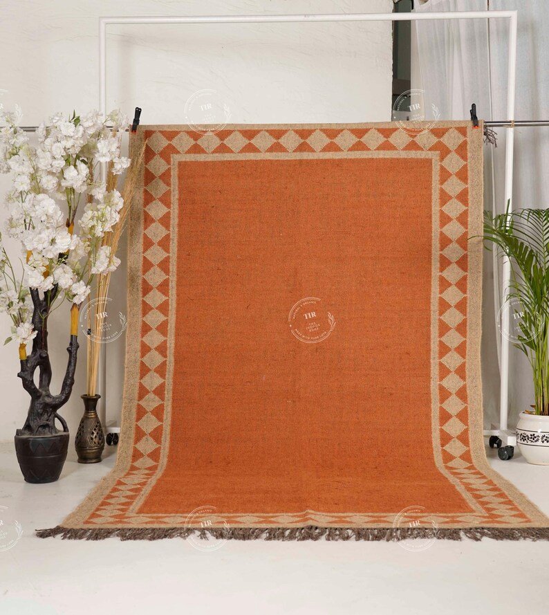 May include: An orange rectangular rug with a geometric diamond pattern border in beige. The rug has a fringed edge and is displayed against a white backdrop. Decorative plants and vases are in the background.