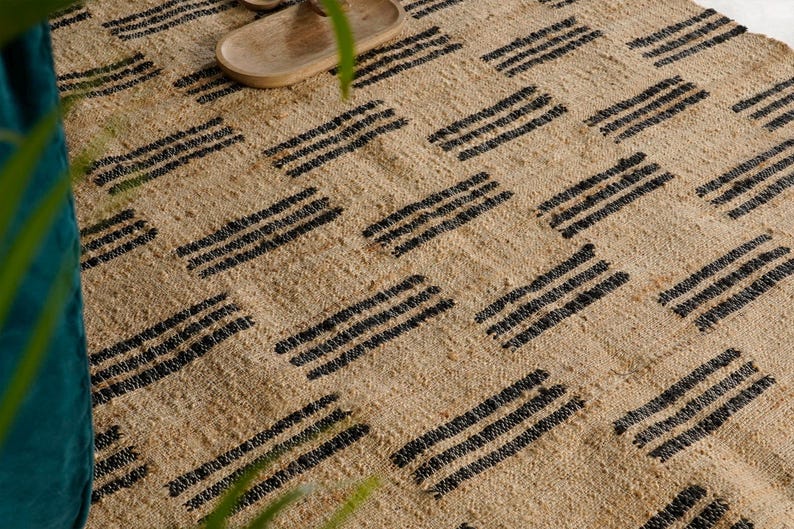 May include: A natural fiber rug with a woven texture and a repeating pattern of black stripes. The rug is beige and has a simple, geometric design. The rug is suitable for indoor use.