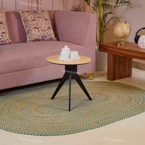 Puede incluir: Una alfombra ovalada trenzada verde y beige est&aacute; sobre un suelo claro. Un sof&aacute; rosa con cojines estampados est&aacute; en el fondo, junto con una mesa de centro de madera y una peque&ntilde;a mesa redonda. Una peque&ntilde;a mesa redonda con base negra y tapa clara est&aacute; en primer plano.