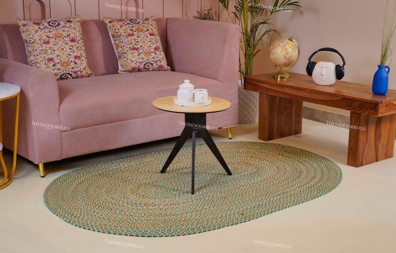 Puede incluir: Una alfombra ovalada trenzada verde y beige est&aacute; sobre un suelo claro. Un sof&aacute; rosa con cojines estampados est&aacute; en el fondo, junto con una mesa de centro de madera y una peque&ntilde;a mesa redonda. Una peque&ntilde;a mesa redonda con base negra y tapa clara est&aacute; en primer plano.