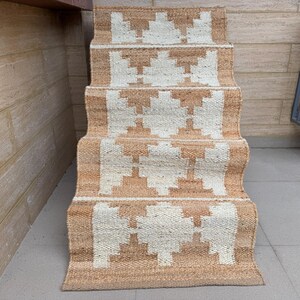 May include: A woven stair runner rug featuring a repeating geometric pattern in natural tan and off-white. The runner is draped over a set of stairs, showcasing a textured, fibrous material with a consistent diamond-like motif across each step.