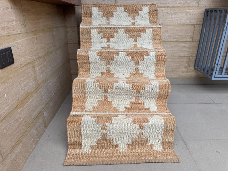May include: A woven stair runner rug featuring a repeating geometric pattern in natural tan and off-white. The runner is draped over a set of stairs, showcasing a textured, fibrous material with a consistent diamond-like motif across each step.