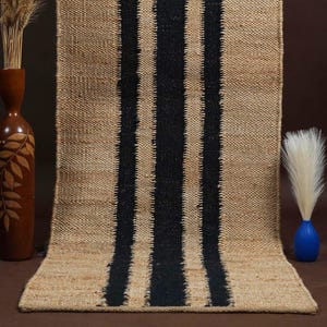 Könnte beinhalten: Ein rechteckiger Jute-Teppich mit drei vertikalen schwarzen Streifen. Der Teppich hat eine natürliche beige Farbe und eine gewebte Textur. Dekorative Gegenstände, darunter eine braune Vase mit Blattgravuren und eine blaue Vase mit weißen Federn, befinden sich im Hintergrund.