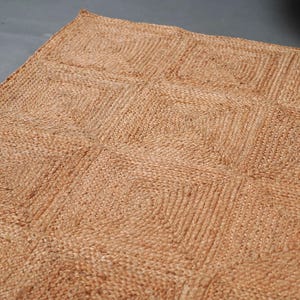 May include: A rectangular, woven jute rug in a natural tan color. The rug features a repeating geometric pattern of squares. The texture of the rug is visible, with the individual fibers creating a tactile appearance. The rug is suitable for indoor use.