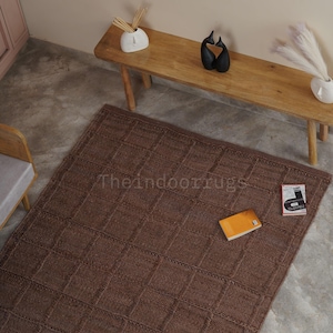 May include: A rectangular brown area rug featuring a textured grid pattern of raised squares. The rug is placed on a concrete floor and displays the text Theindoorrugs printed in the center. An orange book and a small white booklet lie on the rug.