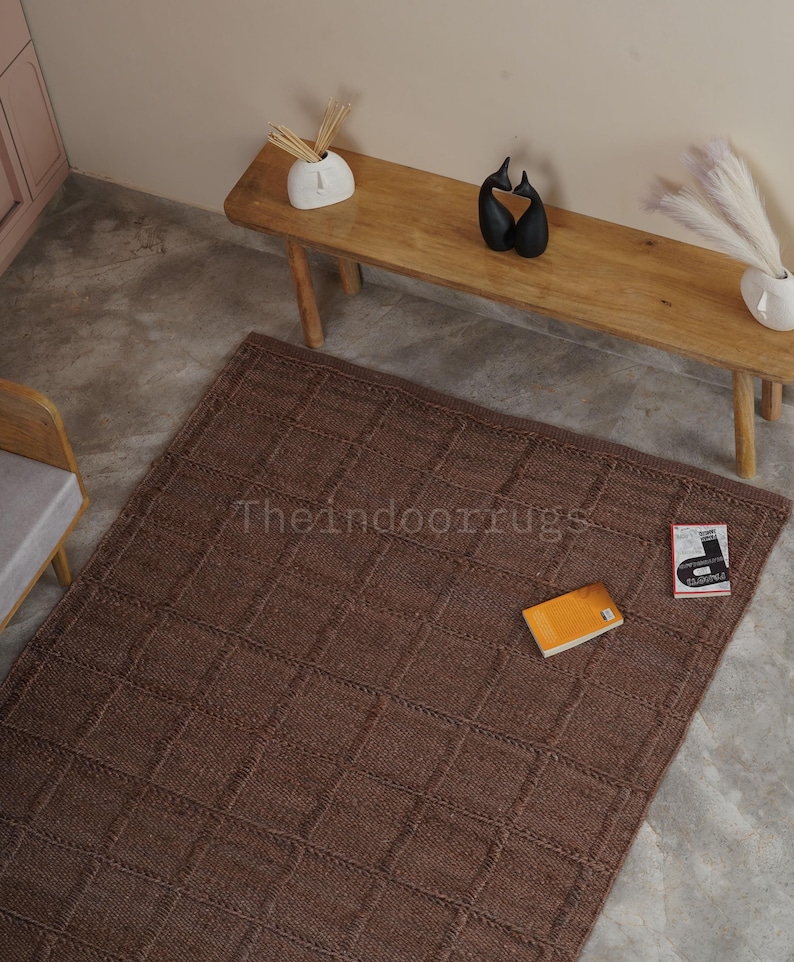 May include: A rectangular brown area rug featuring a textured grid pattern of raised squares. The rug is placed on a concrete floor and displays the text Theindoorrugs printed in the center. An orange book and a small white booklet lie on the rug.