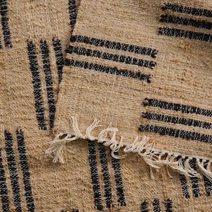 May include: Close-up of a woven rug with a natural beige background and black vertical and horizontal stripe patterns. The rug has a fringed edge. The texture of the rug is visible.