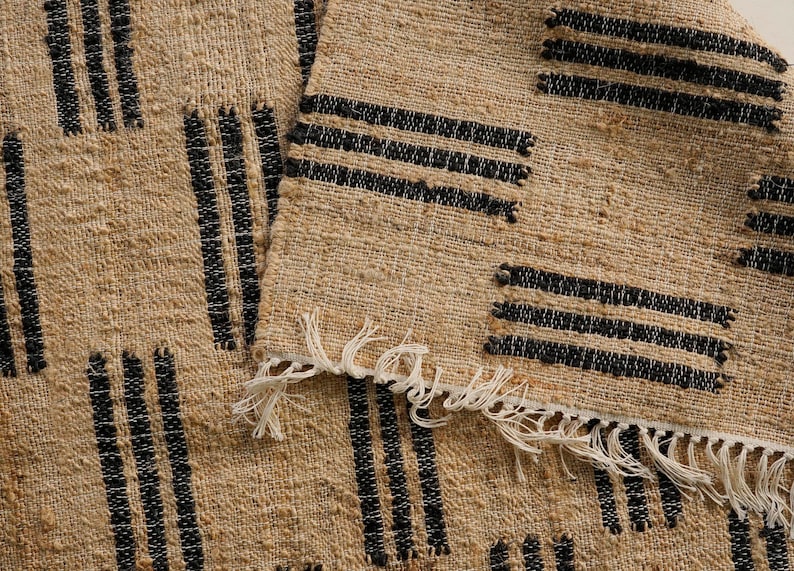 May include: Close-up of a woven rug with a natural beige background and black vertical and horizontal stripe patterns. The rug has a fringed edge. The texture of the rug is visible.