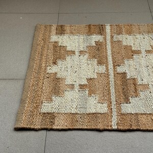 May include: A rectangular woven rug made of natural tan jute fibres featuring a repeating geometric pattern in off-white. The design consists of symmetrical stepped diamond shapes arranged in vertical columns separated by thin vertical white stripes.