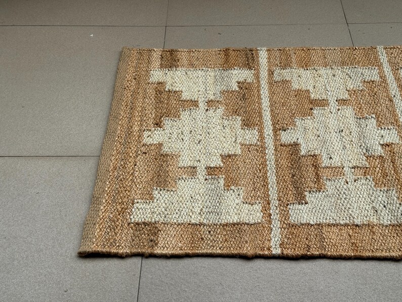 May include: A rectangular woven rug made of natural tan jute fibres featuring a repeating geometric pattern in off-white. The design consists of symmetrical stepped diamond shapes arranged in vertical columns separated by thin vertical white stripes.