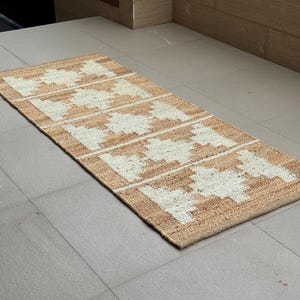 May include: A rectangular runner rug featuring a natural tan jute base with a repeating white geometric diamond pattern. The woven texture is visible throughout the surface, showcasing a series of five distinct diamond shapes aligned horizontally.
