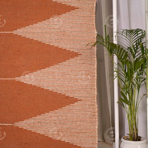 May include: A woven rug with a geometric pattern in shades of orange and white. The design features large, repeating chevrons. The rug is made of a textured material, and the edges are neatly finished. The rug is displayed near a window with a potted plant.
