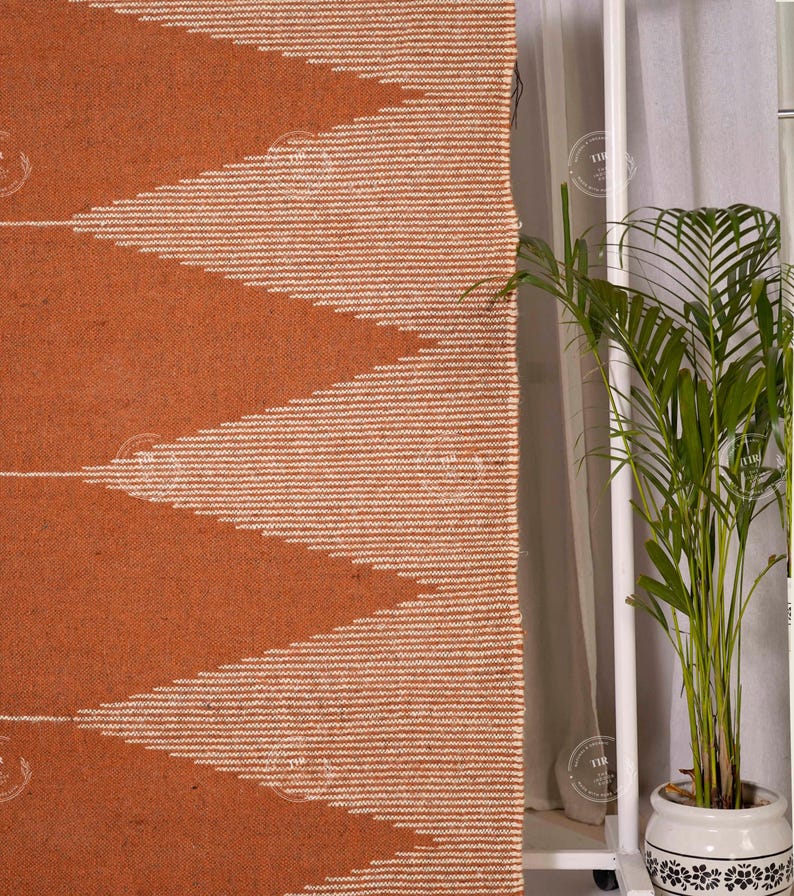May include: A woven rug with a geometric pattern in shades of orange and white. The design features large, repeating chevrons. The rug is made of a textured material, and the edges are neatly finished. The rug is displayed near a window with a potted plant.