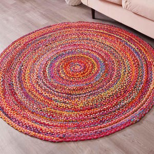 K&ouml;nnte beinhalten: Ein runder geflochtener Teppich mit einem Spiralmuster. Der Teppich zeigt eine lebendige Mischung aus Farben, darunter Rot, Rosa, Orange, Gelb und Blau. Der Teppich liegt auf einem hellen Holzboden.