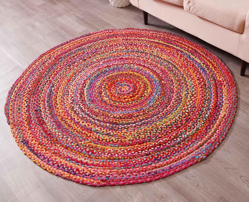 K&ouml;nnte beinhalten: Ein runder geflochtener Teppich mit einem Spiralmuster. Der Teppich zeigt eine lebendige Mischung aus Farben, darunter Rot, Rosa, Orange, Gelb und Blau. Der Teppich liegt auf einem hellen Holzboden.
