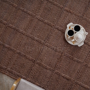 May include: A brown woven area rug featuring a grid pattern of raised, textured lines. A white ceramic tea set consisting of a small teapot and two cups filled with dark liquid sits on a matching white tray placed on the surface of the rug.