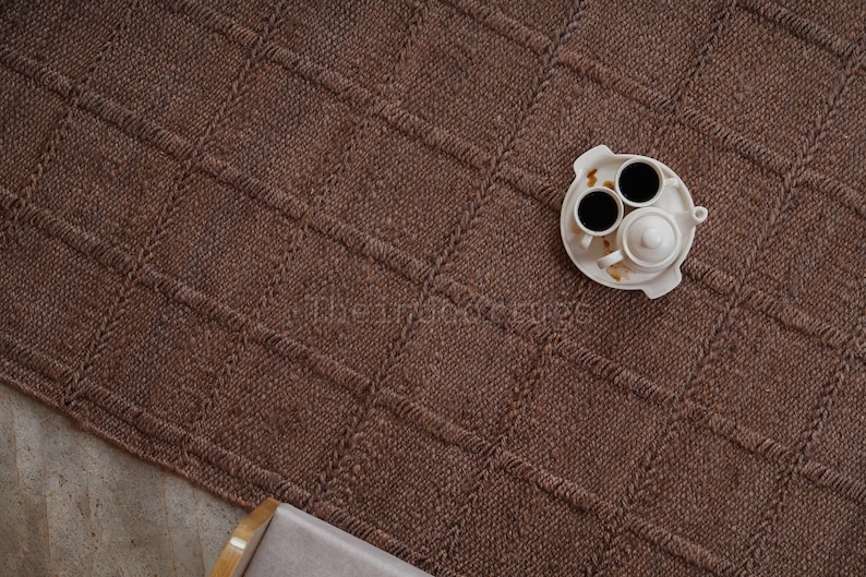 May include: A brown woven area rug featuring a grid pattern of raised, textured lines. A white ceramic tea set consisting of a small teapot and two cups filled with dark liquid sits on a matching white tray placed on the surface of the rug.