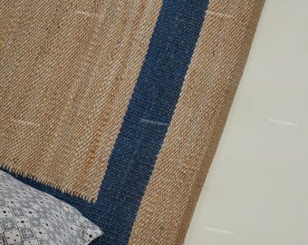 Puede incluir: Una alfombra de fibra natural con textura tejida, con una ancha franja azul marino en el centro. La alfombra está parcialmente enrollada, revelando su construcción. Se puede ver una almohada estampada en la esquina.